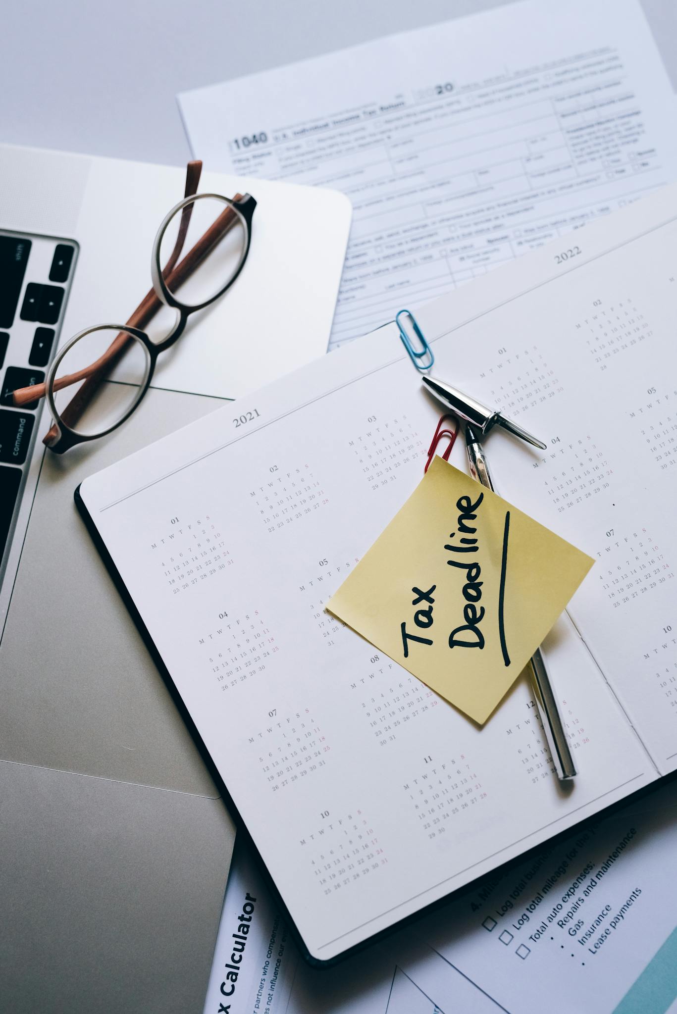 Calendar with tax deadline sticky note on desk with documents, glasses, and laptop.